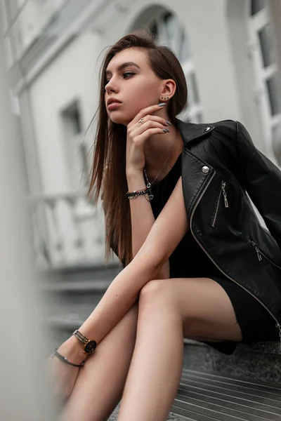 Jolie jeune femme mince à la mode dans une robe élégante noire dans une veste en cuir vintage se détend assis sur un escalier en pierre à l'extérieur. Tendance moderne fille mignonne modèle reposant dans la ville. Style jeunesse . — Photo