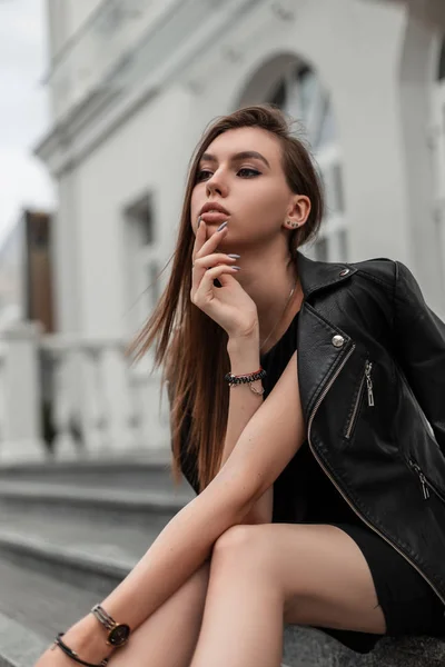 Urban szczupła piękna młoda kobieta w czarnej krótkiej modnej sukience w stylowej skórzanej kurtce siedzi na kamiennych schodach w pobliżu zabytkowego białego budynku w mieście.Modne cute girl relaksuje się na ulicy — Zdjęcie stockowe