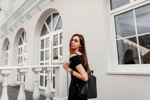 Gioiosa bella giovane donna modello in un elegante abito nero con un elegante zaino nero in pelle viaggia per la città. La ragazza alla moda felice gode una passeggiata su una strada. Street moda giovanile . — Foto Stock