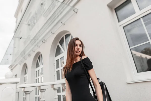 Slim αρκετά χαρούμενη νεαρή γυναίκα μοντέλο σε ένα χαριτωμένο χαμόγελο σε ένα κομψό μαύρο φόρεμα με γυμνούς ώμους με ένα δερμάτινο μαύρο μοντέρνο σακίδιο απολαμβάνει μια βόλτα στην πόλη. Ευτυχισμένο κορίτσι ξεκουράζεται έξω. — Φωτογραφία Αρχείου