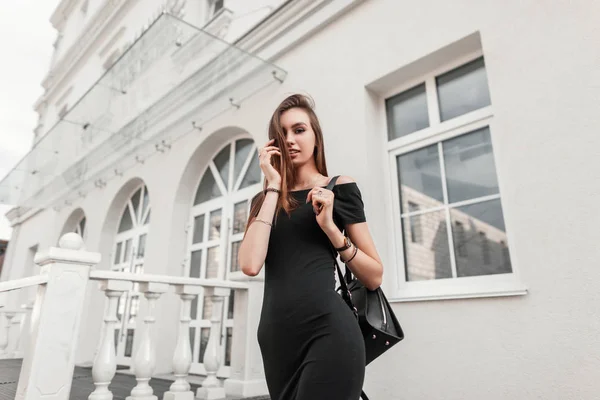 Jolie jolie jeune femme mannequin de mode avec les cheveux bruns dans une robe noire à la mode avec un sac à dos élégant posant dans la ville près d'un bâtiment blanc moderne. Belle fille urbaine sur une promenade . — Photo