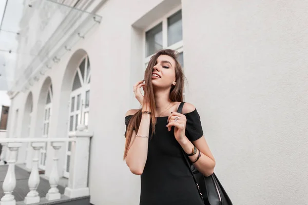 レザーファッショナブルなバックパック付きのスタイリッシュな黒のドレスでかわいい笑顔の幸せな若い女性は、暖かい秋の日に街を歩くことを楽しんでいます。魅力的な流行の女の子まっすぐ髪屋外. — ストック写真