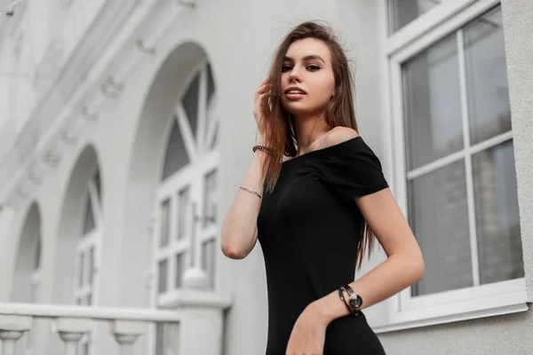 Lindo modelo de mujer joven europea en ropa negra de moda endereza el cabello cerca de un edificio blanco. Hermosa chica bonita de moda en un vestido elegante con hombros desnudos al aire libre en un día de primavera . — Foto de Stock