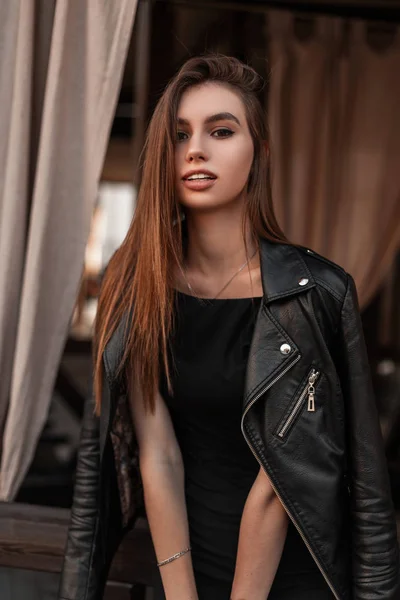 Bonito bela jovem mulher em uma jaqueta preta de couro na moda em um vestido elegante preto está descansando em um café vintage. Menina atraente gosta do fim de semana na cidade . — Fotografia de Stock
