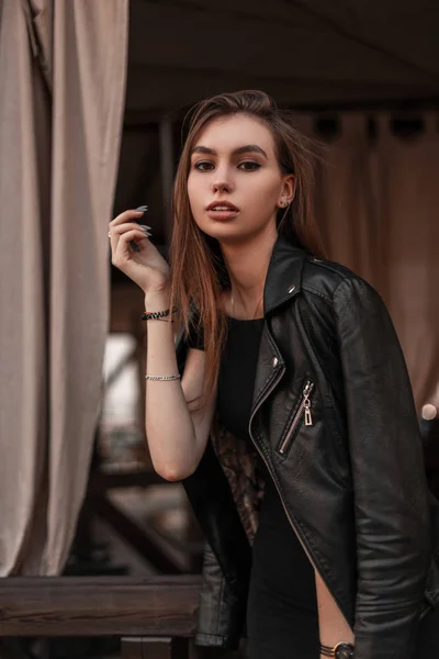 Moderno europeu bela jovem mulher em uma jaqueta de couro elegante preto em um vestido preto elegante está descansando em um café vintage. Modelo de moda menina muito atraente gosta do fim de semana na cidade . — Fotografia de Stock