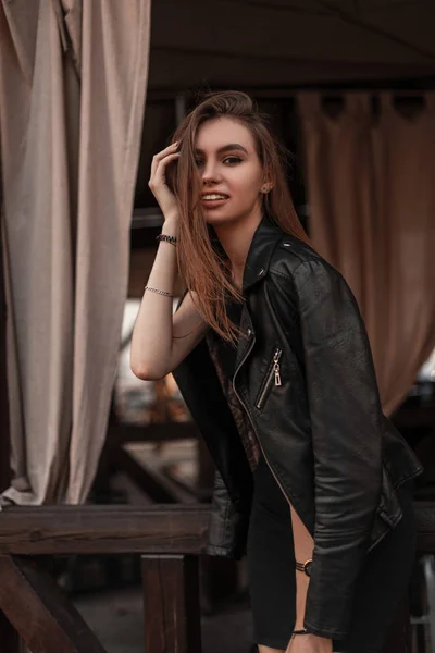 Feliz hermosa mujer joven en una chaqueta de cuero negro de moda en un vestido elegante negro está descansando en un café vintage. Chica alegre disfruta del fin de semana en la ciudad . —  Fotos de Stock