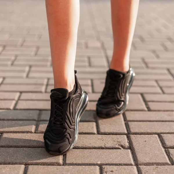 Moderne jonge vrouw met slanke mooie benen in modieuze zwarte sneakers loopt door de straat. Stijlvolle sportieve damesschoenen. Zomer stijl. Close-up van vrouwelijke benen met schoenen. — Stockfoto