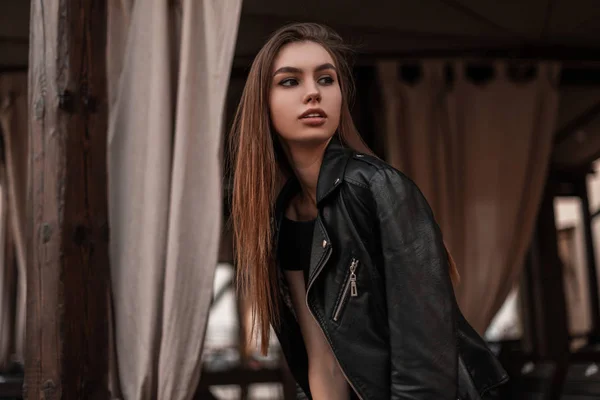 Moderno europeu bela jovem mulher em uma jaqueta de couro elegante preto em um vestido preto elegante está descansando em um café vintage. Bonita modelo de moda menina urbana gosta do fim de semana na cidade . — Fotografia de Stock