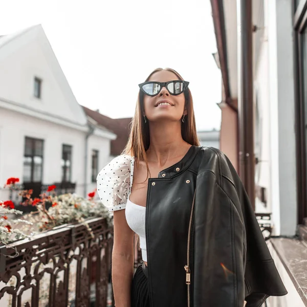 Веселая молодая хипстерша с положительной улыбкой в модных солнцезащитных очках в черной куртке наслаждается отдыхом стоя на винтажном балконе в осенний солнечный день. Радостная девушка отдыхает на открытом воздухе . — стоковое фото