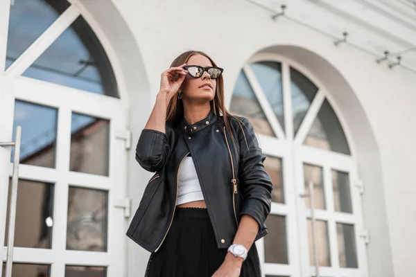 Mulher hipster jovem na moda em endireita óculos de sol na moda. Modelo elegante menina urbana em roupas sazonais posando na cidade perto de um edifício branco vintage em um dia quente de outono. Estilo de juventude . — Fotografia de Stock