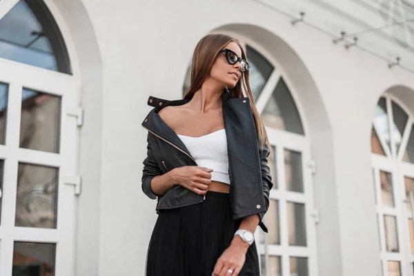 Junge hübsche trendige Hipsterfrau in stylischer schwarzer Lederjacke, elegantem Oberteil und modischer Sonnenbrille, die in der Nähe eines historischen Gebäudes in der Stadt posiert. europäisches Mädchen Modell ruht sich auf der Straße aus. — Stockfoto