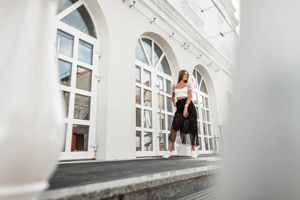 Ευτυχισμένη νέα μοντέρνα γυναίκα σε μια κομψή μαύρη φούστα σε ένα κομψό τοπ με λευκά sneakers που ποζάρουν κοντά σε ένα vintage κτίριο με παράθυρα στην πόλη. Χαριτωμένο χαρούμενο κορίτσι με όμορφο χαμόγελο σε εξωτερικούς χώρους. — Φωτογραφία Αρχείου