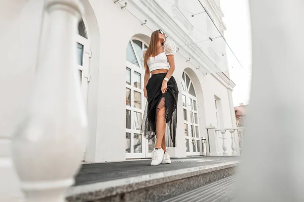 Позитивная молодая хипстерша в модной летней черно-белой одежде в стильной кожаной обуви в модных солнцезащитных очках позирует рядом с белым винтажным зданием на улице. Счастливая девушка гуляет по городу . — стоковое фото