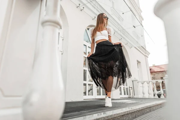 Ganska glad ung kvinna i en snygg svart kjol i trendiga solglasögon i en topp i fashionabla skor poserar utomhus nära en modern byggnad i staden. Attraktiv glad hipster flicka njuter av en promenad. — Stockfoto