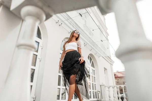 Elegante linda mujer hipster joven con una hermosa sonrisa en ropa glamorosa con estilo en gafas de sol pasea por la ciudad cerca de un edificio blanco vintage. Atractiva modelo de chica positiva. Estilo de verano . — Foto de Stock