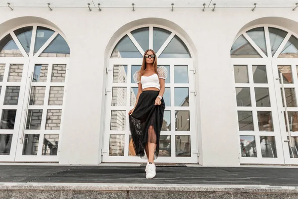 Hermosa joven hipster con gafas de sol en una elegante falda larga negra en un top de moda en zapatillas de cuero posando cerca de un edificio vintage en la ciudad. Atractiva chica feliz está descansando al aire libre . —  Fotos de Stock