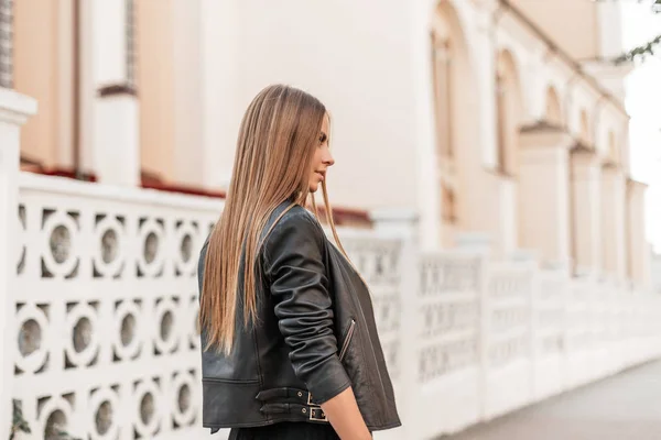 Elegante jovem mulher moderna com um longo cabelo castanho na moda jaqueta de couro preto posando em uma rua na cidade de cerca branca perto do vintage. Menina atraente na moda gosta de relaxar ao ar livre . — Fotografia de Stock