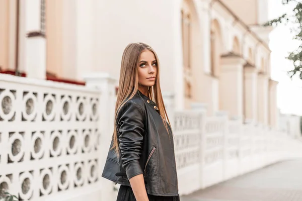 Jolie jolie jeune femme modèle avec beau maquillage naturel dans une veste vintage en cuir noir avec les cheveux bruns posant à l'extérieur près d'une clôture blanche. Joyeuse fille mignonne se promène autour de la ville . — Photo