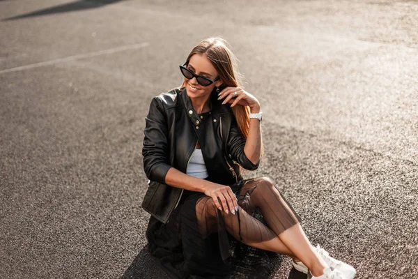Joyful jonge hipster vrouw in modieuze jas in een rok in een sneakers in stijlvolle zonnebril zit op het asfalt op een zonnige herfstdag. Positief meisje model met een leuke glimlach is rusten op een zon. — Stockfoto
