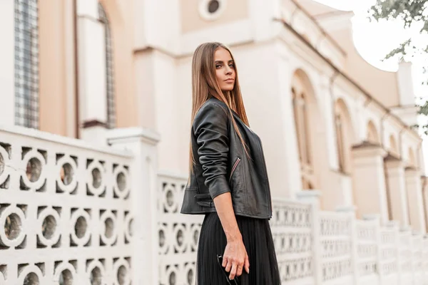 Bastante elegante modelo de mujer joven en ropa elegante y elegante está de pie cerca de una cerca blanca vintage en la ciudad en un cálido día de otoño. Chica hermosa moderna está descansando en la calle. Estilo . — Foto de Stock