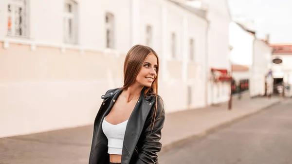 Modell av en attraktiv glad ung kvinna med vacker naturlig makeup i en vintage skinnjacka i en snygg vit blus med brunt hår färdas på en gata. Glädjande söt flicka går på stan. — Stockfoto