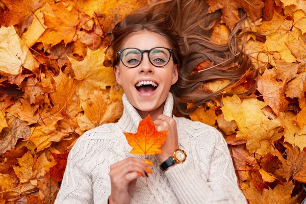 Смішна молода хіпстерка в модному в'язаному светрі в стильних окулярах з помаранчевим листом в руках виражає сюрприз, лежачи на осінньому листі в лісі. Позитивна дівчина модель відпочинку . — стокове фото