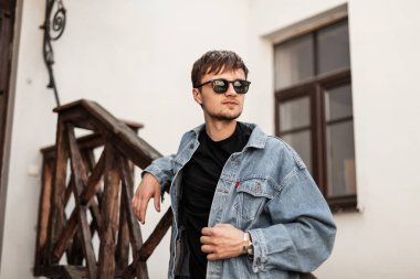Havalı saç stili, klasik güneş gözlüklü, mavi kot ceketli, beyaz bir binanın yanında sokakta poz veren şık bir genç. Çekici trend hipster adam. Bahar stili.