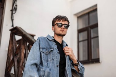 Son moda saç stili, şık güneş gözlüklü, klasik kot ceketli, şehirde beyaz bir binanın yanında poz veren, modaya uygun bir genç adam. Çekici modern hippi adam. Bahar stili.