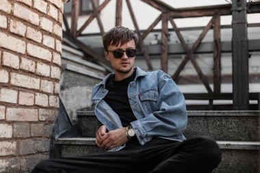 Genç Avrupalı stil sahibi genç adam modaya uygun gençlik kıyafetleri içinde modaya uygun siyah güneş gözlükleriyle şehirde bir tuğla duvarın yanında rahatlıyor. Çekici adam hipster sokağın tahta merdivenlerinde dinleniyor..