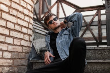 Güneş gözlüklü, siyah pantolonlu, mavi kot ceketli hippi genç adam caddedeki klasik tuğla duvarın yanındaki merdivenlerde dinleniyor. Çekici stil sahibi adam şehirde rahatlıyor. Bahar stili.