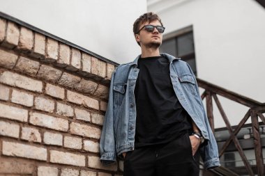 Modaya uygun mavi kot ceketli, güneş gözlüklü havalı genç hippi adam sokaktaki klasik tuğla duvarın yanında duruyor. Dışarıda şık bir saç stili olan yakışıklı bir adam. Moda bahar erkek giyimi.