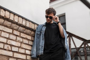 Modern Amerikalı genç hippi, tuğla bir binanın yanında şık güneş gözlüğü takıyor. Şehirde poz veren şık kot elbiseli çekici adam modeli. Bahar modası. Moda erkek giyim.