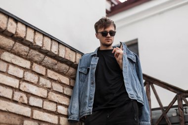 Şık kentsel erkek modeli modaya uygun gençlik kotları içinde güneş gözlükleriyle tuğla duvarın yanında poz veriyor. Genç yakışıklı hippi adam şehirdeki eski bir binanın yanında dinleniyor. Bahar modası.