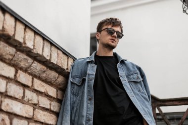 Modaya uygun mavi kot ceketli, güneş gözlüklü modern modaya uygun genç hippi şehirdeki eski bir tuğla duvarın yanında duruyor. Havalı saç stili olan çekici bir moda adamı. Erkek giyim.