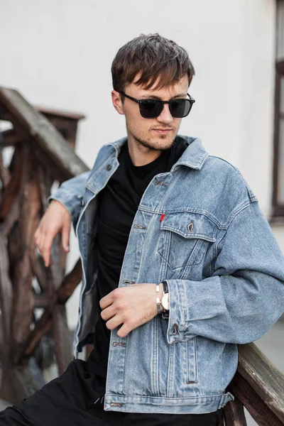 Ung stilig man hipster i en fashionabel blå denim jacka i snygga solglasögon i svarta jeans vilar i staden nära vintage byggnad. Attraktiv kille kopplar av utomhus i staden. Herrkläder. — Stockfoto