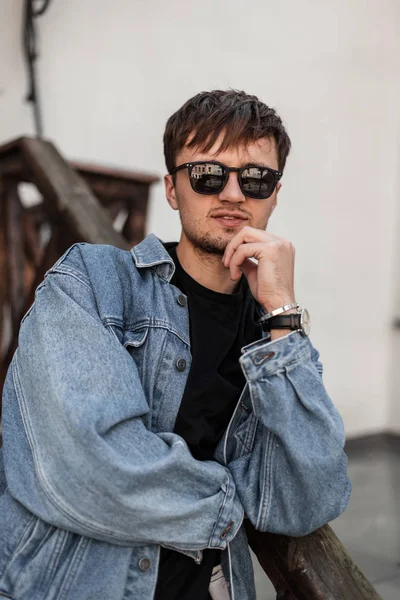 Amerykański, brutalny hipster w modnej, jeansowej kurtce w okularach przeciwsłonecznych z brodą spoczywa w pobliżu drewnianej poręczy w mieście w wiosenny dzień. Atrakcyjny miejski stylowy facet cieszy się wakacjami — Zdjęcie stockowe