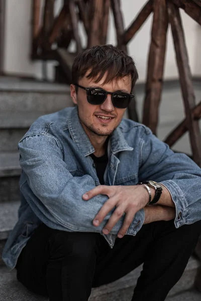 Happy cool młody człowiek hipster w stylowe niebieskie dżinsowe ubrania w modnych okularach przeciwsłonecznych siedzi na zabytkowych schodach w mieście w ciepły wiosenny dzień. Radosny atrakcyjny facet z uroczym uśmiechem odpoczywa na świeżym powietrzu. — Zdjęcie stockowe