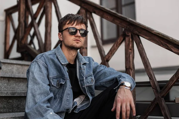 Joven hombre hipster guapo en una chaqueta de mezclilla de moda en pantalones negros en gafas de sol oscuras con estilo relajarse sentado en una escalera vintage en la ciudad en un día de primavera.Hombre moderno disfruta de un fin de semana al aire libre — Foto de Stock