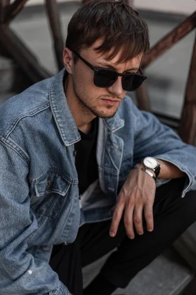 Fashionabla unga man hipster i eleganta denim kläder trendiga i en svart solglasögon sitter på en vintage trappa i staden på en vårdag. Attraktiv cool kille vilar utomhus. Trendiga herrkläder. — Stockfoto