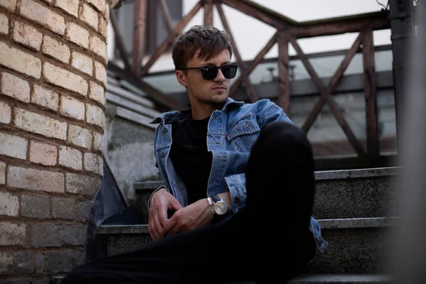 Cool joven hipster en ropa de mezclilla azul con estilo en gafas de sol de moda se sienta en una escalera vintage cerca de la pared de ladrillo en la ciudad en un cálido día de primavera. El muchacho a la moda con el peinado descansa al aire libre . — Foto de Stock