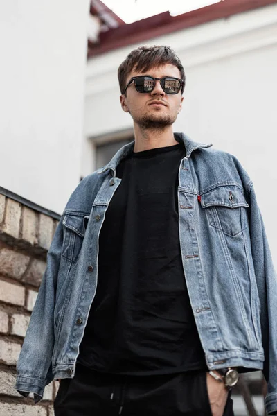 Brutalnie przystojny młody mężczyzna model z brodą z fryzurą w modnej odzieży stoi w pobliżu zabytkowej cegły ściany w mieście. Fajny facet hipster w okularach przeciwsłonecznych w dżinsowej kurtce spoczywa na zewnątrz. — Zdjęcie stockowe