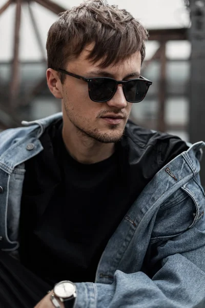 Joven guapo americano guapo con una barba en gafas de sol de moda en una chaqueta de mezclilla azul con estilo en una camiseta se encuentra en la calle en un día de primavera. Un hipster urbano al aire libre. Estilo de calle . — Foto de Stock