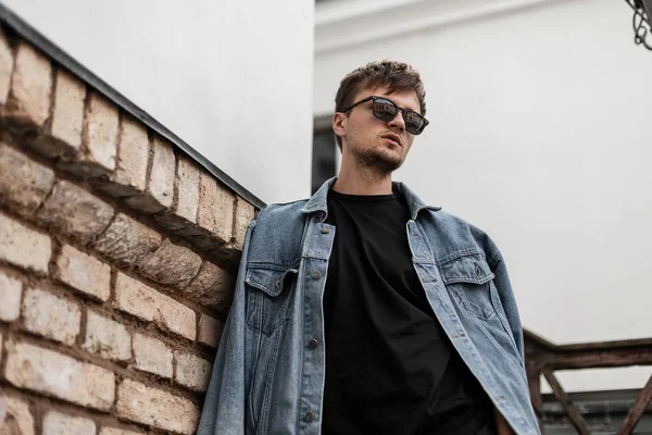 Moderno hombre hipster joven de moda en una chaqueta de mezclilla azul de moda en gafas de sol se encuentra cerca de una pared de ladrillo vintage en la ciudad. Muchacho de moda atractiva con un peinado elegante al aire libre. Prendas masculinas . — Foto de Stock