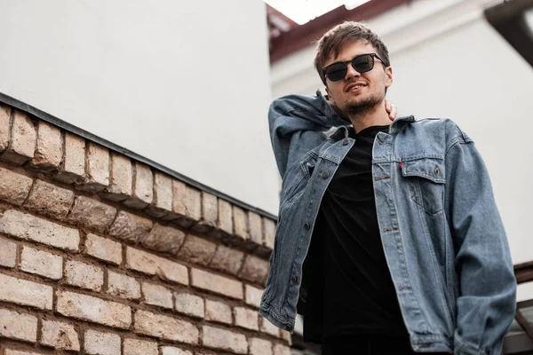 Bonito jovem americano hipster em uma jaqueta jeans azul vintage em uma camiseta preta em óculos de sol elegantes posando perto de um edifício vintage na cidade. Atraente cara positivo com um sorriso bonito ao ar livre . — Fotografia de Stock