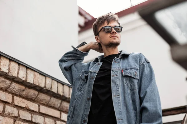 El joven hipster a la moda endereza el peinado de estilo. Guapo chico moderno en ropa vaquera de moda en gafas de sol posando en un día de primavera cerca de una pared de ladrillo vintage en la calle. Estilo juvenil . — Foto de Stock