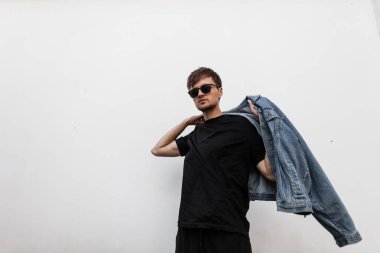 Çekici genç adam kot ceket giyiyor. Yakışıklı, sakallı, güneş gözlüklü, şık gençlik kıyafetleriyle şehirde beyaz bir vintage binanın yanında poz veren modern bir hippi. Bahar modası.