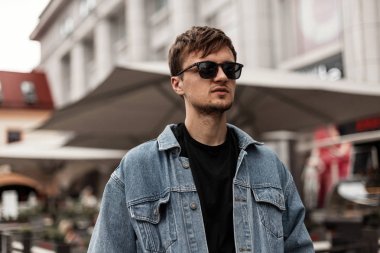 Şık mavi kot ceketli, güneş gözlüklü havalı genç hippi şehirde sokakta duruyor. Havalı bir saç stili olan yakışıklı adam dışarıda dinleniyor. Moda bahar erkek giyimi.