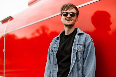 Sakallı, genç, neşeli bir hippi. Tişörtü içinde, modaya uygun bir kot ceketle şehirde Red Metal bir binanın yanında poz veriyor. Sokaktaki mutlu adam. Erkek giyim.