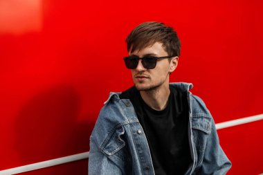 Modaya uygun mavi kot ceketli, şık güneş gözlüklü caddedeki kırmızı metal duvarın yanında sakallı, son moda genç hippi. Çekici erkek modeli şehirde bir yaz günü dinleniyor..