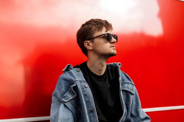 Sakallı, şık bir saç stili olan, şık bir kot ceketli, güneş gözlüklü, şehirde Red metal bir binanın yanında poz veren çekici bir hippi. Sokaktaki havalı adam.